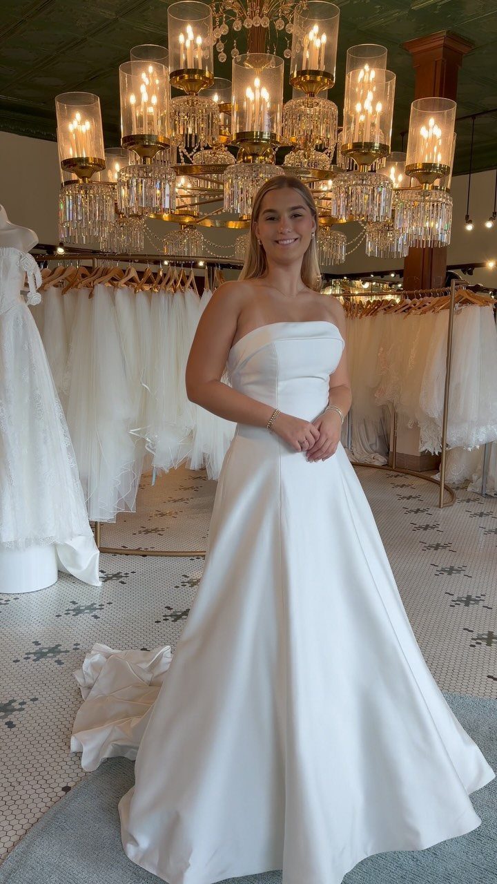 2026 Wedding Dresses: 12 Bridal Styles to Know Right Now 3 A woman smiling while wearing a strapless white satin wedding gown in a bridal boutique.