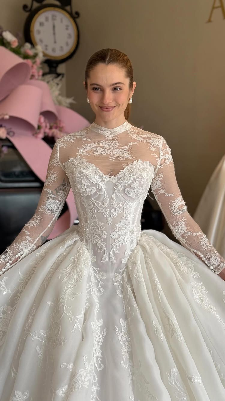 2026 Wedding Dresses: 12 Bridal Styles to Know Right Now 1 Woman smiling while wearing a white long-sleeved wedding dress with intricate lace detailing.