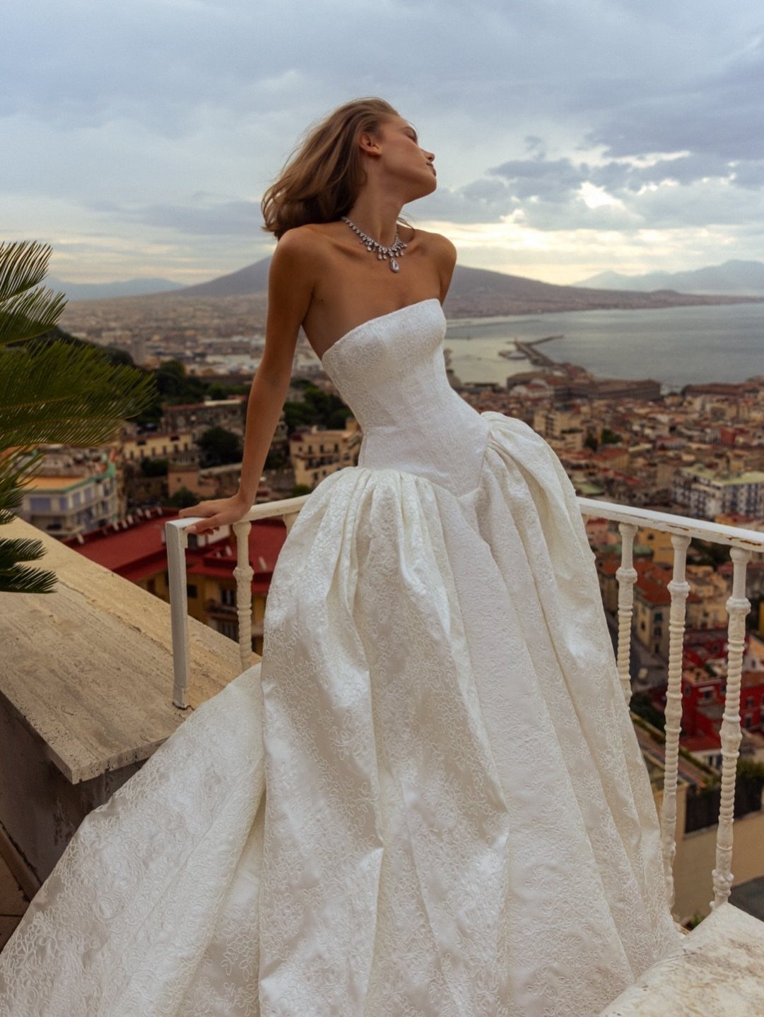 2026 Wedding Dresses: 12 Bridal Styles to Know Right Now 12 Woman in a strapless white wedding gown standing on a balcony overlooking a coastal city.
