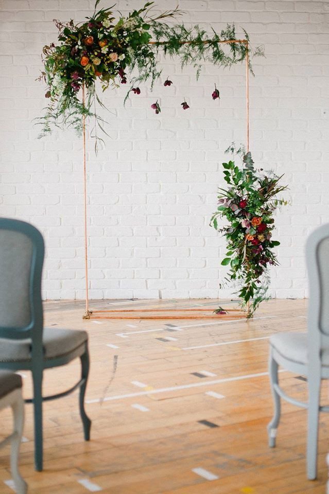 Alternative Wedding Arch: 8 Beautiful Styles for Your Ceremony 3 Copper metal rectangular arch decorated with green foliage and colorful flowers against a white brick wall.