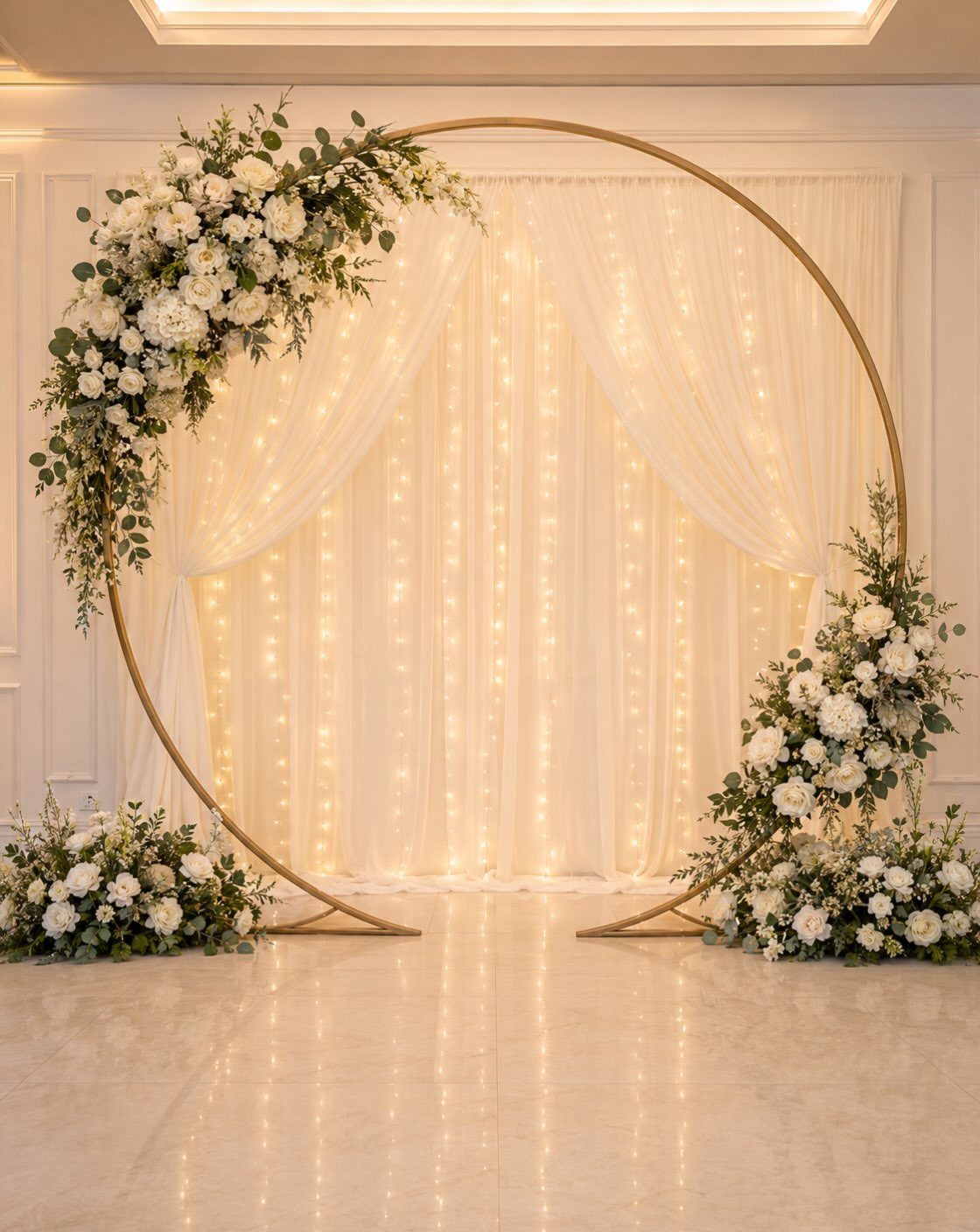 Alternative Wedding Arch: 8 Beautiful Styles for Your Ceremony 4 A large circular gold metal wedding arch decorated with white flowers and greenery against lit sheer curtains.