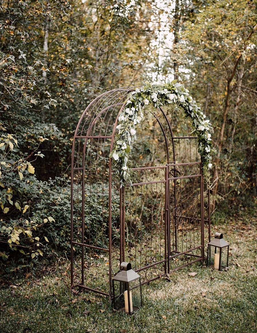 Alternative Wedding Arch: 8 Beautiful Styles for Your Ceremony 6 A metal garden arch adorned with white flowers stands in a forest setting.