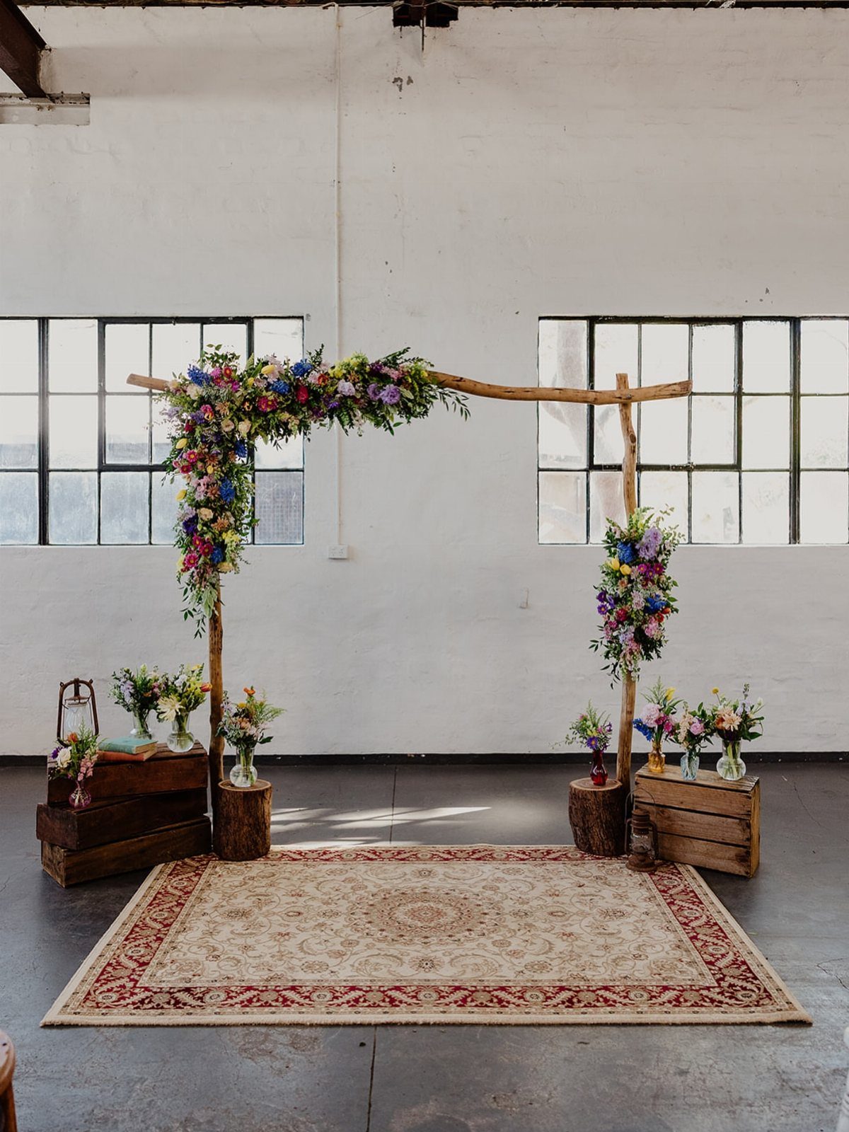 Alternative Wedding Arch: 8 Beautiful Styles for Your Ceremony 2 Wooden branch arch decorated with colorful wildflowers standing over a patterned rug in an industrial space.