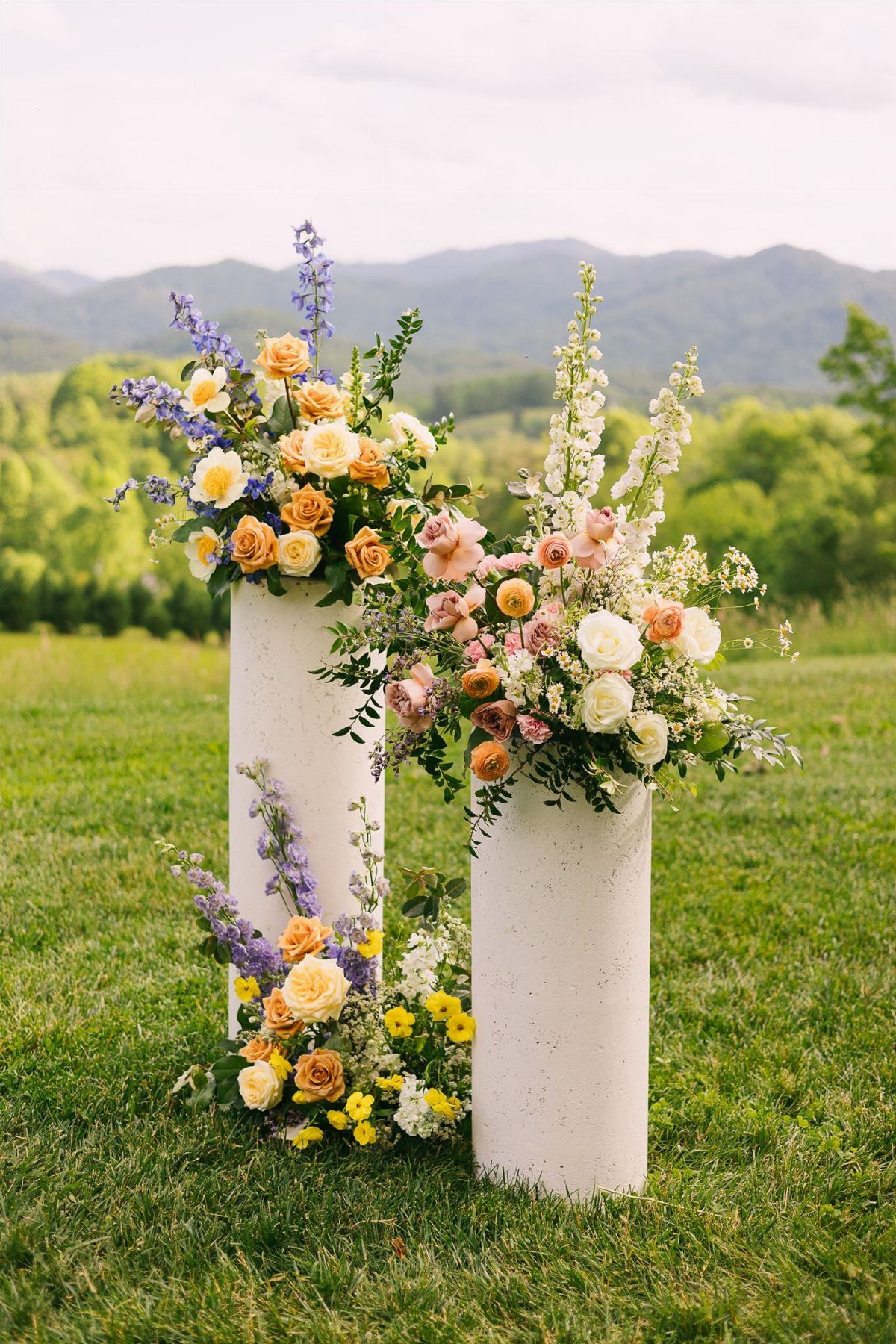 Alternative Wedding Arch: 8 Beautiful Styles for Your Ceremony 5 White cylindrical pillars adorned with colorful peach and purple wildflower arrangements in a grassy field.
