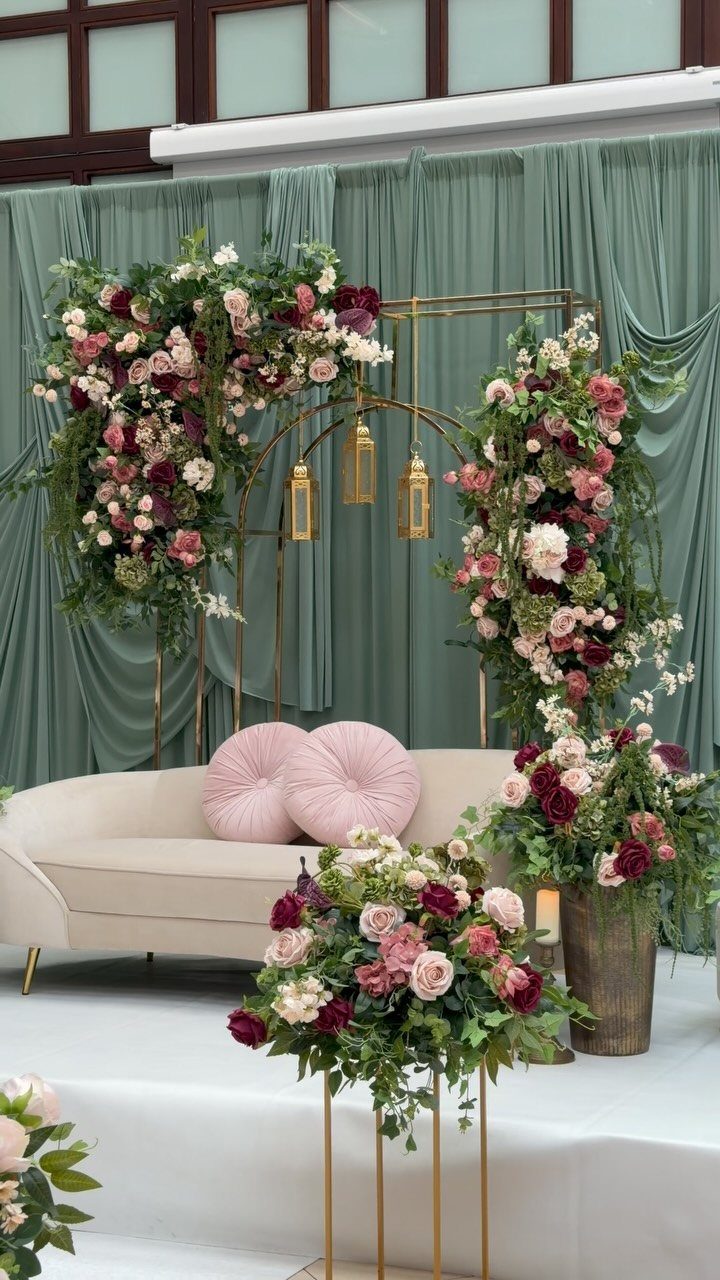 Blush Wedding Colors: 9 Palettes That Work at Actual Weddings 1 Blush and burgundy floral arrangements decorating a gold arch and vases against sage green curtains.
