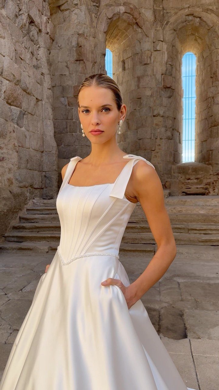 Woman wearing a white satin wedding dress with bows on the shoulder straps against stone ruins.