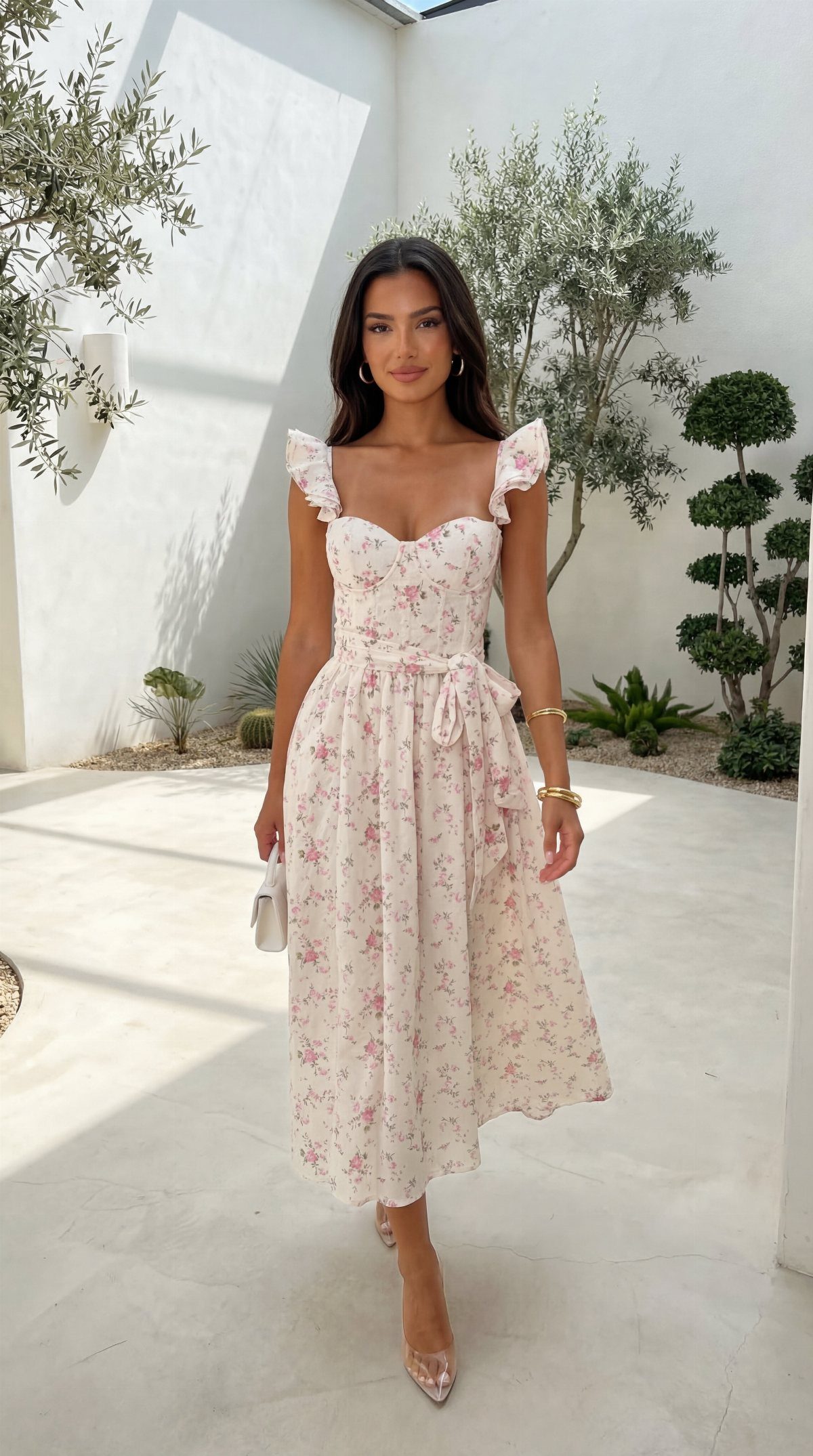 A woman posing in a white floral midi dress with ruffled sleeves in a sunny courtyard.