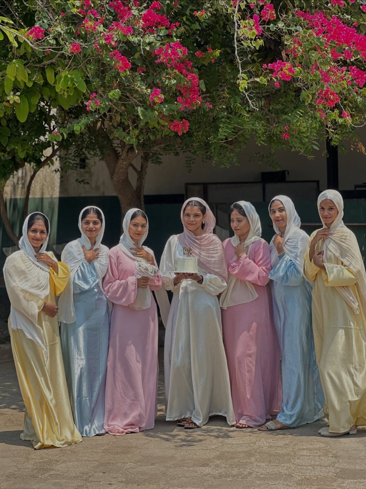 Seven women in pastel flowing dresses and headscarves standing under a pink flowering tree.