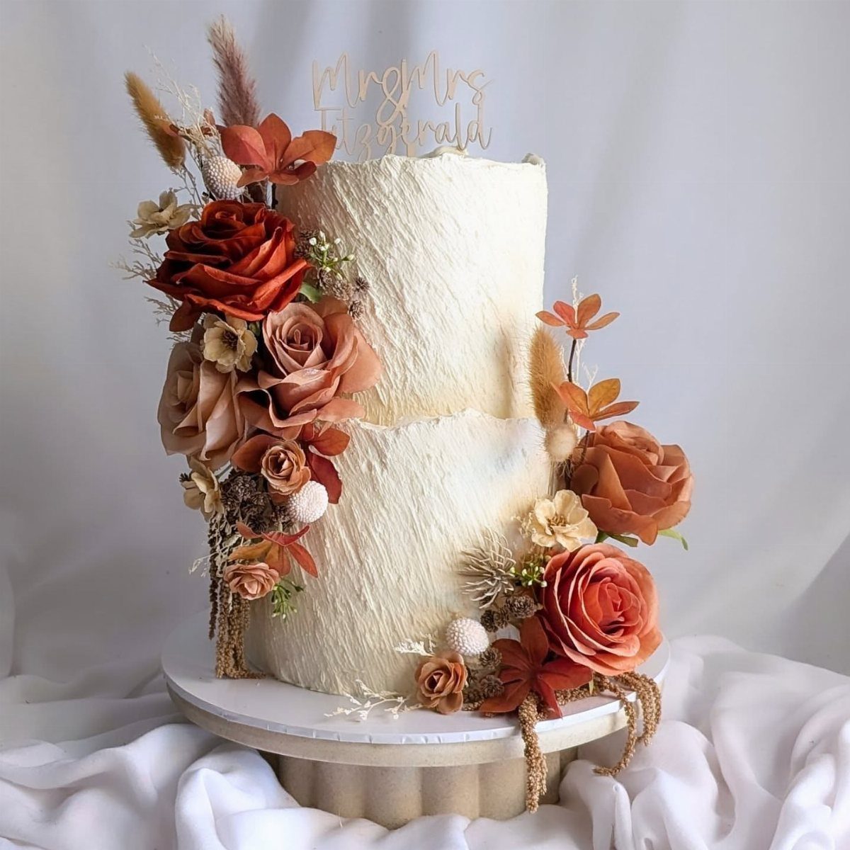 Two-tier white textured wedding cake decorated with burnt orange roses and dried autumnal florals.