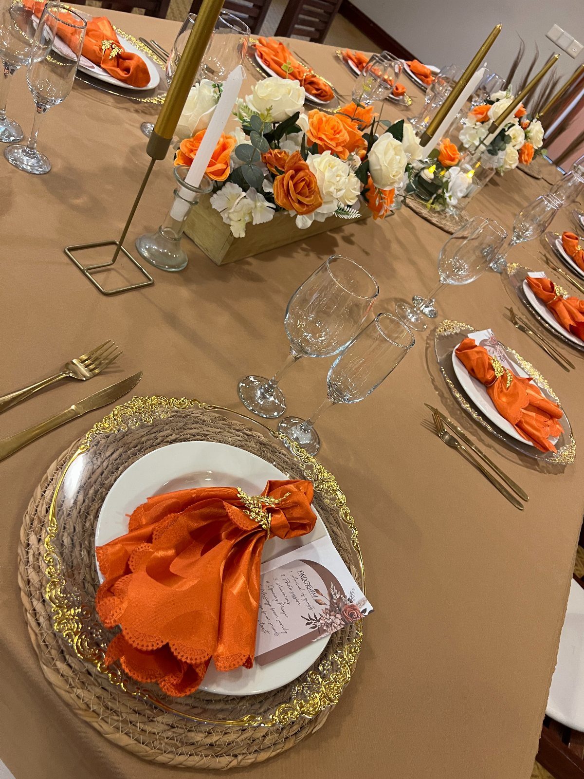 A wedding table set with burnt orange napkins, gold cutlery, and orange and white floral centerpieces.