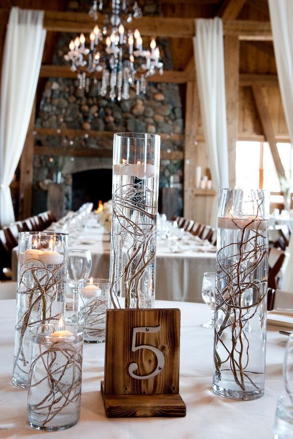 Tall glass cylinder vases containing submerged willow branches and floating white candles on a wedding table.