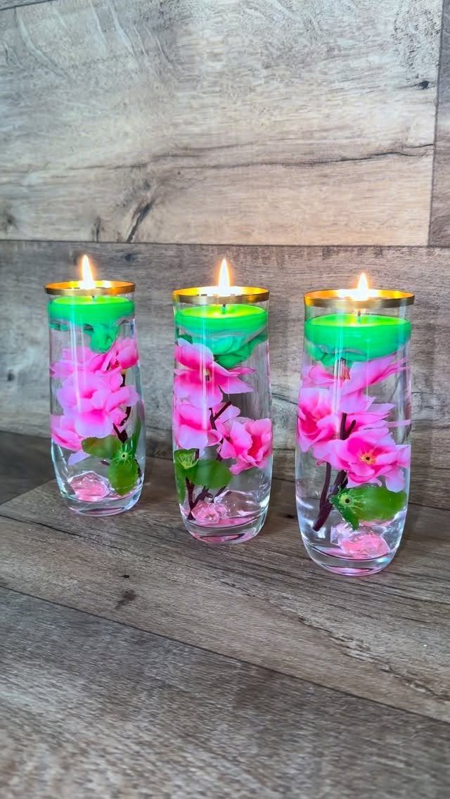 Three glass cylinder vases with pink flowers and floating green candles on a wooden surface.