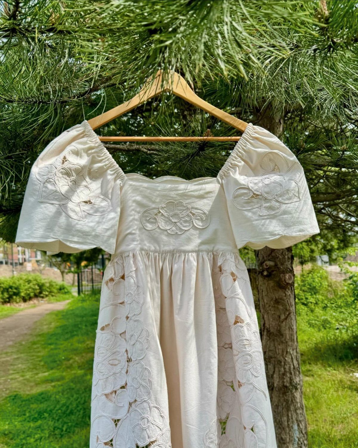 6 Classy Vintage Wedding Dress Finds That Hold Up 5 A cream vintage wedding dress with floral embroidery hanging on a wooden hanger against a pine tree.