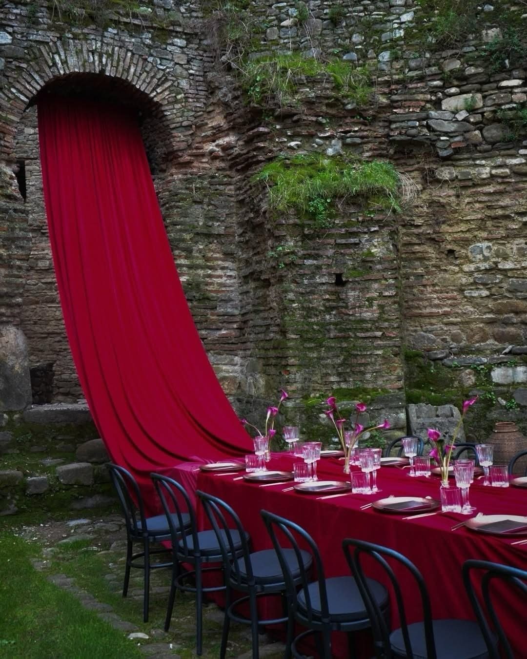 Dark Red Wedding Colors: 20 Ideas for Mood and Palette 5 A long table with burgundy linens and glassware set against a rustic stone wall background.