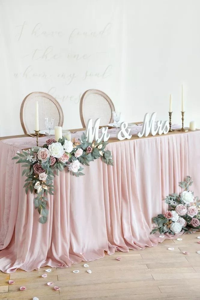 A wedding sweetheart table with dusty rose satin cloth and sage green floral arrangements.