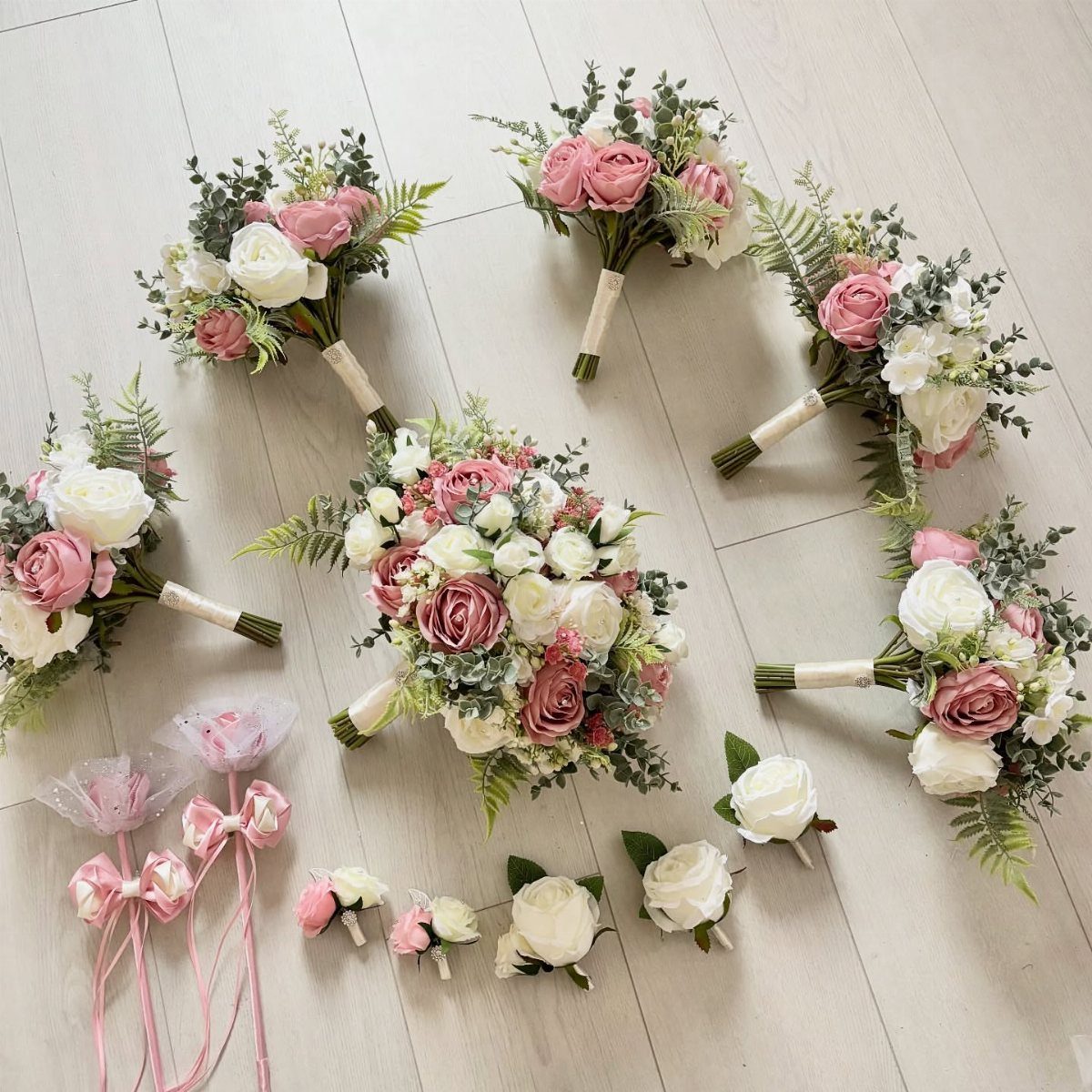 Dusty rose and white rose bouquets with sage green greenery arranged on a light wooden floor.