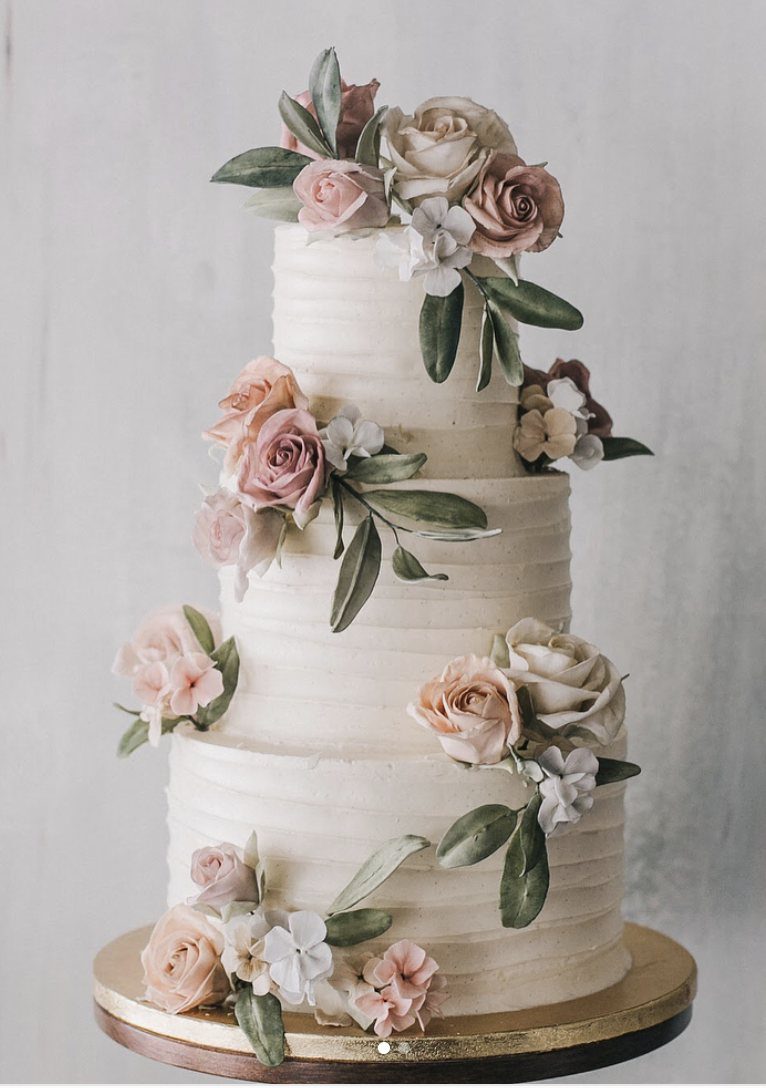 A three-tier white wedding cake decorated with dusty rose flowers and sage green leaves.