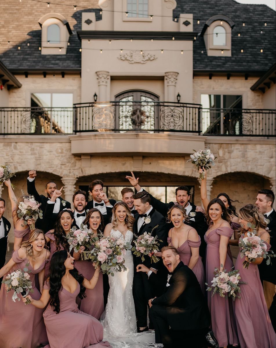 Wedding party in dusty rose dresses and black suits posing in front of a large stone mansion.