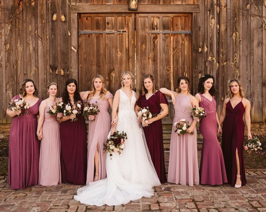 Bridesmaids in dusty rose and burgundy dresses standing with a bride in front of a wooden barn.