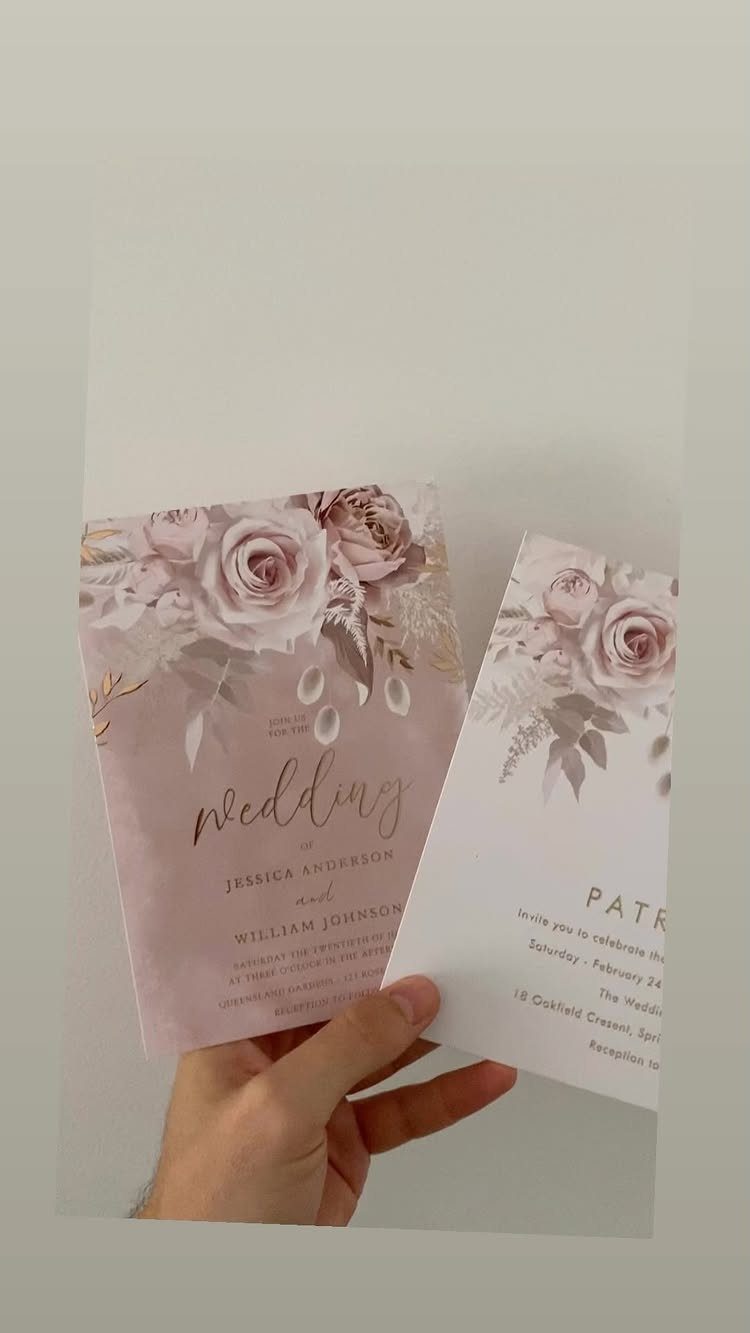 A hand holding two dusty rose wedding invitations decorated with floral rose illustrations and elegant script.