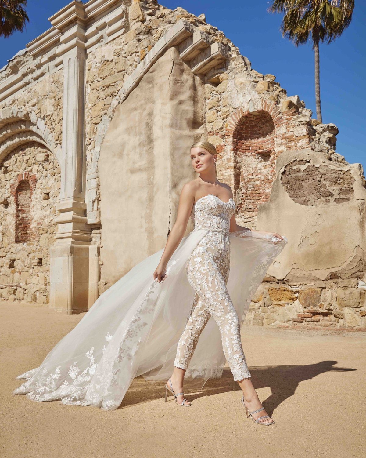 A woman wearing a white lace bridal jumpsuit with a long detachable tulle train.