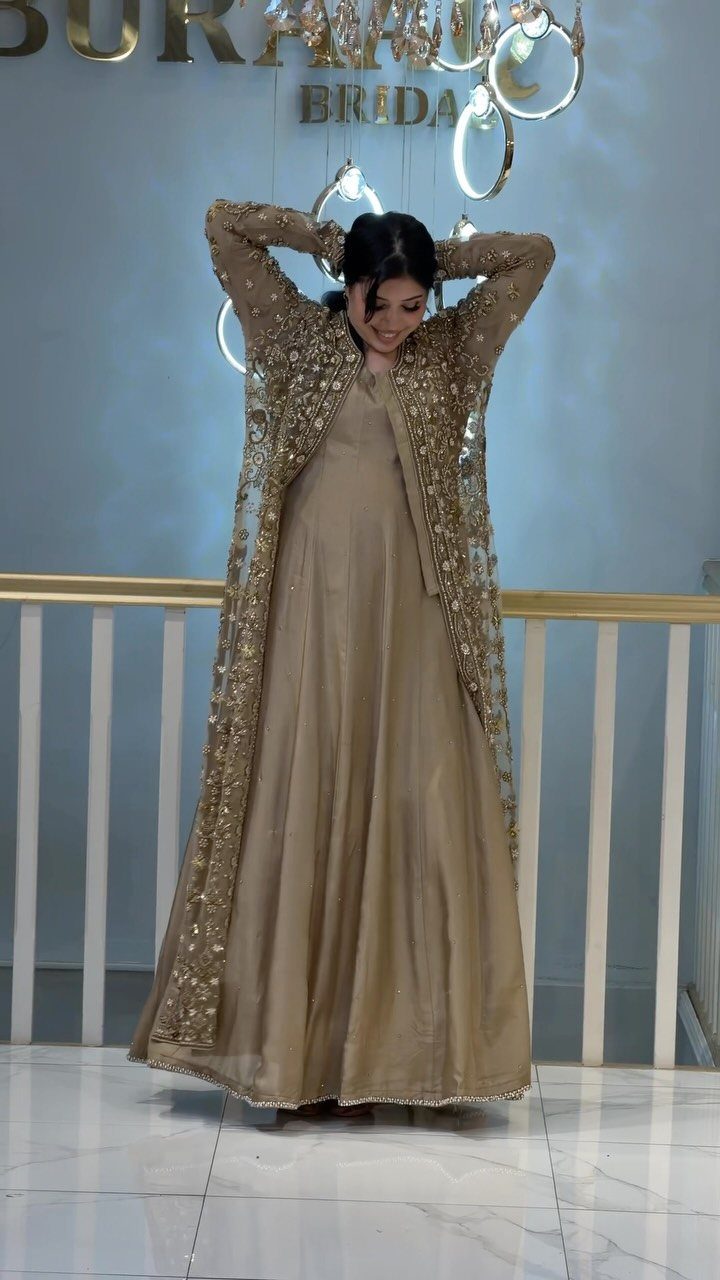 Woman in a champagne gown and gold embroidered sheer jacket standing against a blue wall.