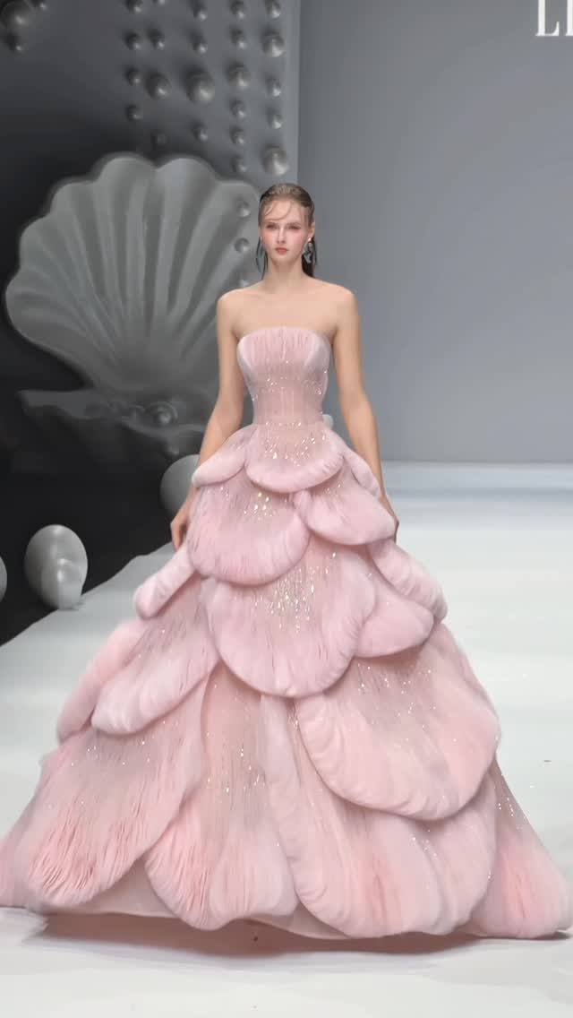 A woman in a voluminous, petal-layered pink strapless gown walking on a runway.