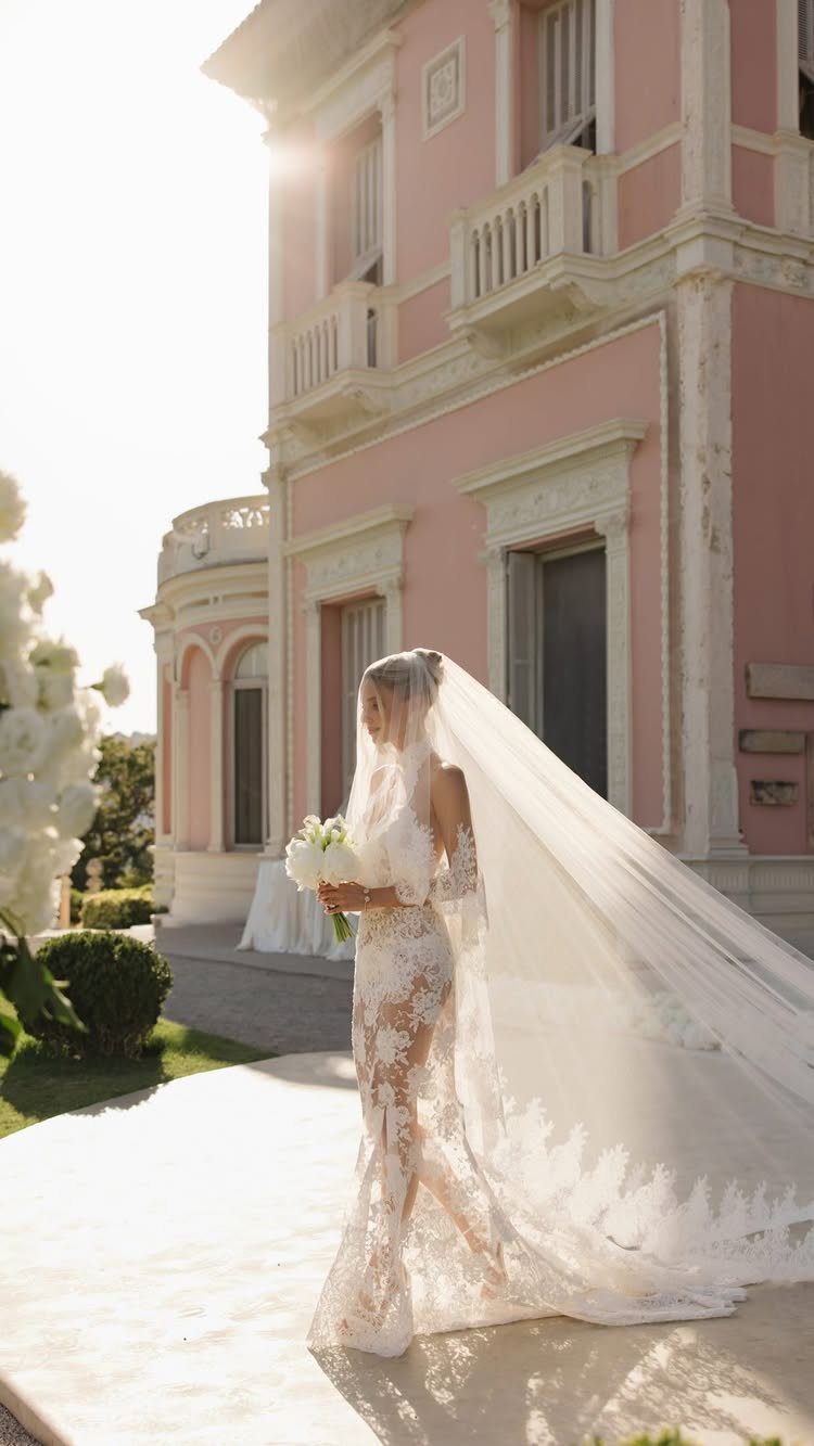 A bride wearing a sheer lace wedding dress and long veil standing before a pink villa.