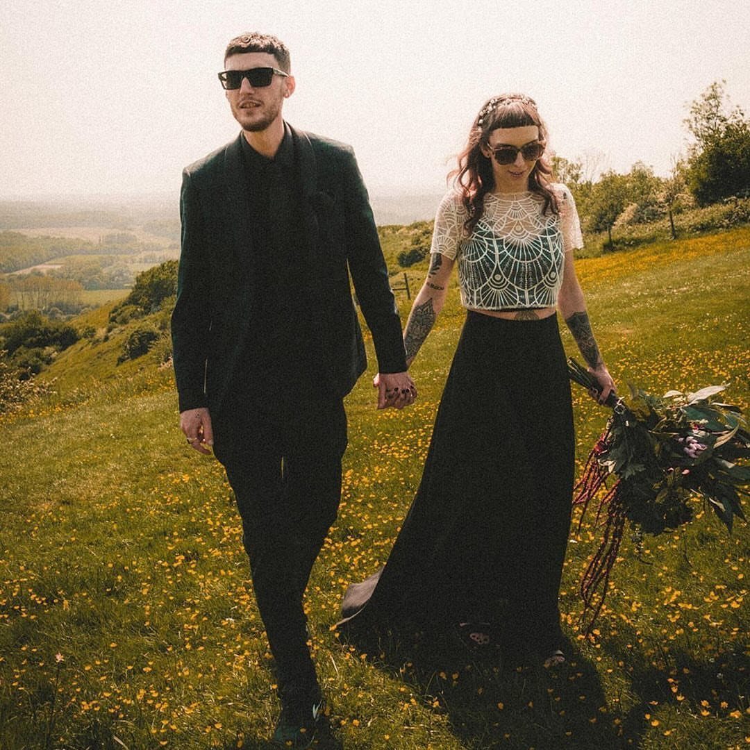 A couple walking through a sunny meadow, the woman wearing a black two-piece bridal outfit.
