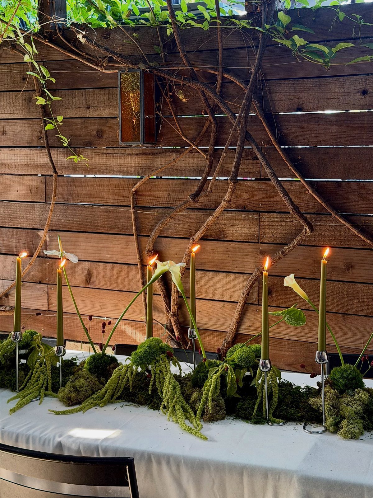 Green taper candles and fresh moss arranged on a white tablecloth against a wooden fence.