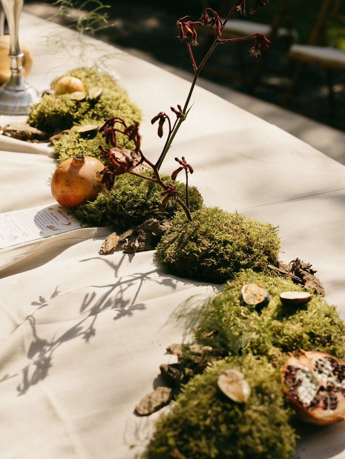 Green moss runner decorated with pomegranates and bark on a white tablecloth.