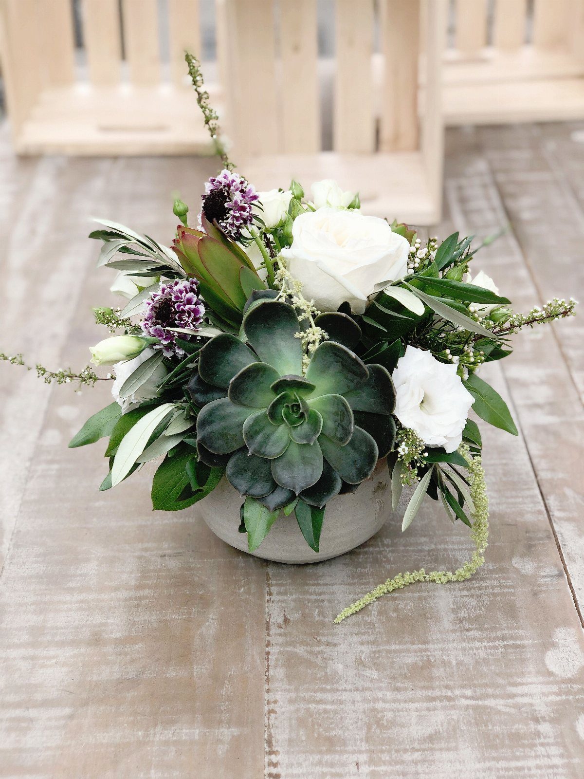 A centerpiece arrangement featuring green succulents, white roses, and purple flowers in a white bowl.