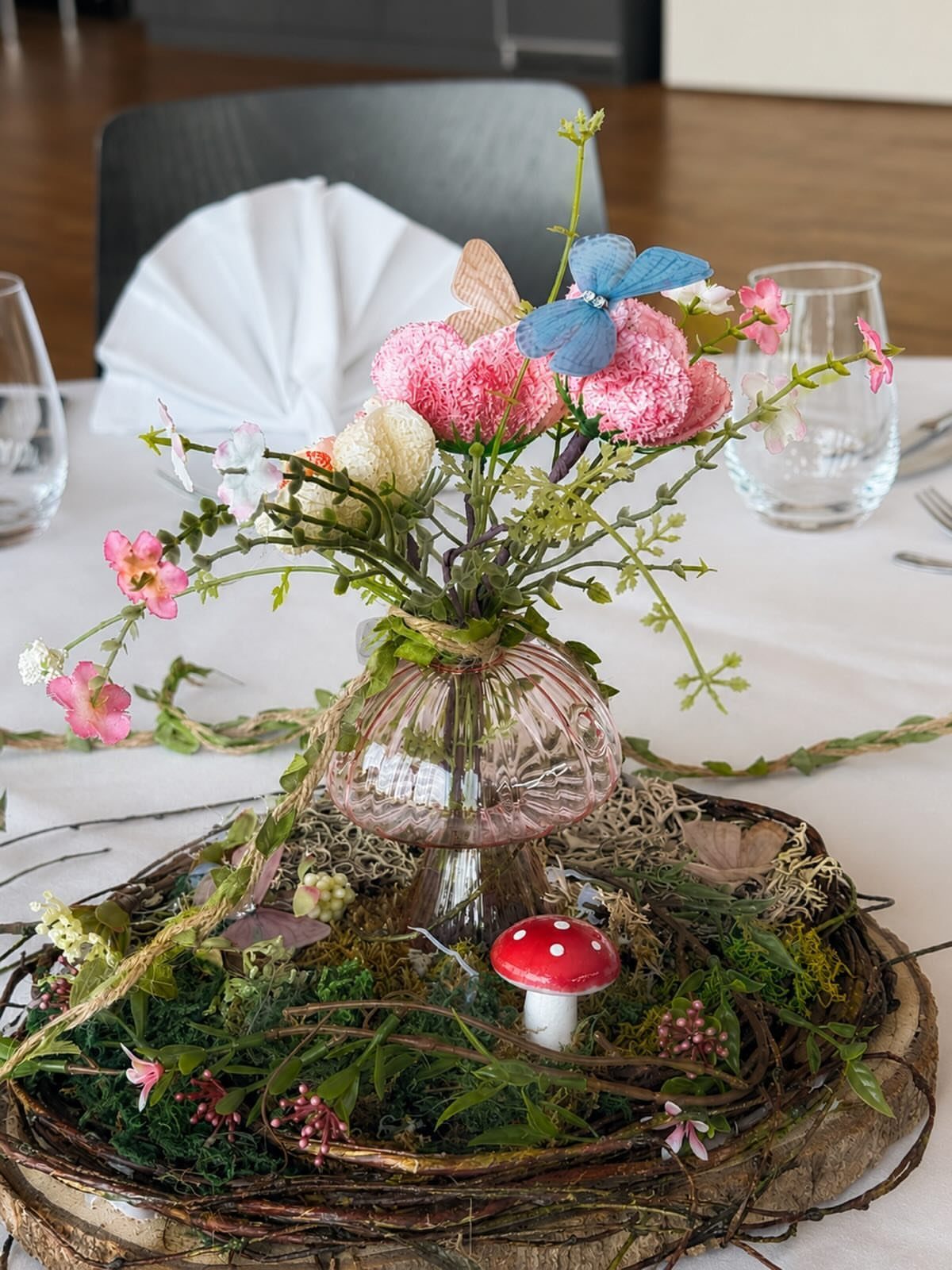 A pink glass vase with wildflowers and butterflies sits atop a mossy wood slice base.
