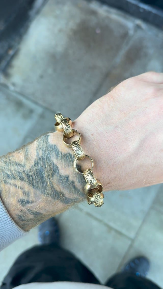A gold Belcher chain bracelet worn on a tattooed person's wrist outdoors.