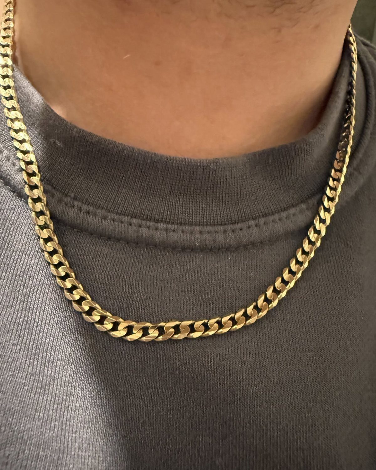 A thick gold curb chain necklace worn by a person over a grey t-shirt.
