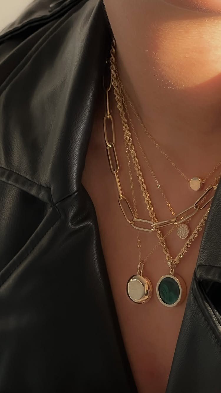 Layered gold necklaces with a paperclip chain and circular pendants worn over a black leather jacket.