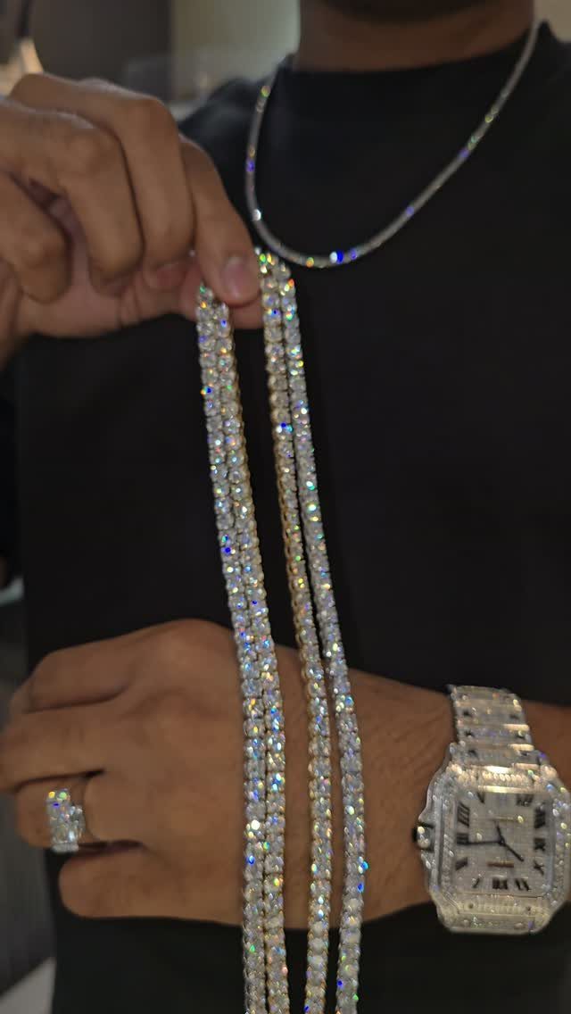 A person holding multiple sparkling tennis chains against a black shirt while wearing a diamond watch.