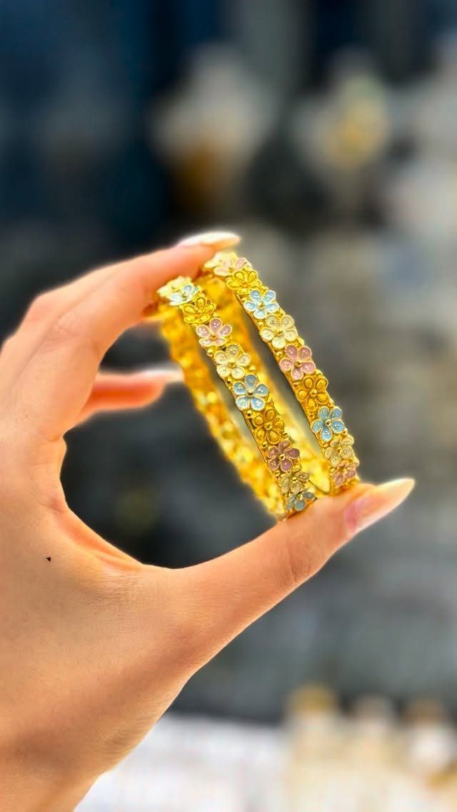 A hand holding two gold bangles adorned with colorful enamel floral patterns.