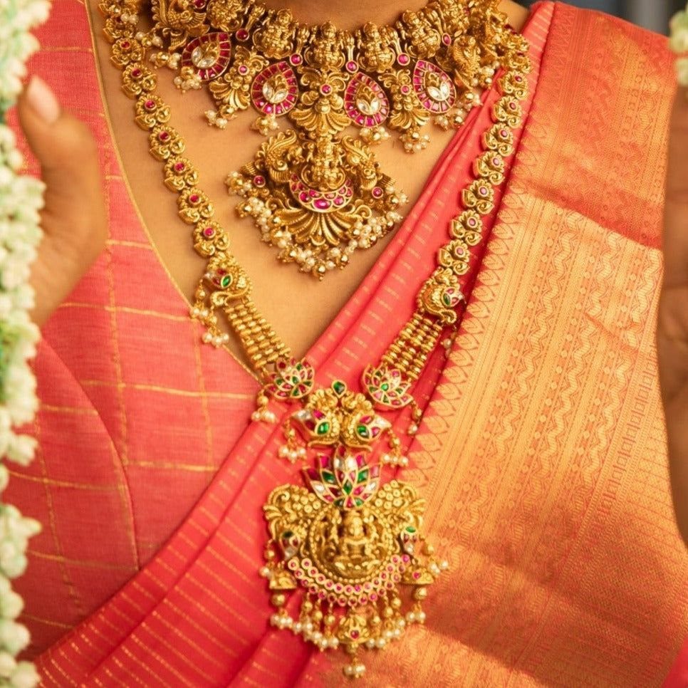 Gold long haram necklace with deity motifs worn over an orange silk saree.