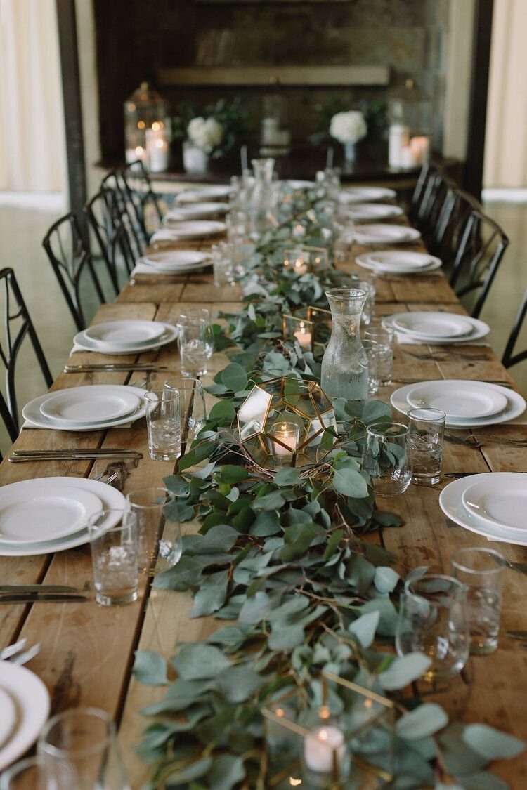 A long wooden table decorated with a green eucalyptus garland and lit candles in glass holders.