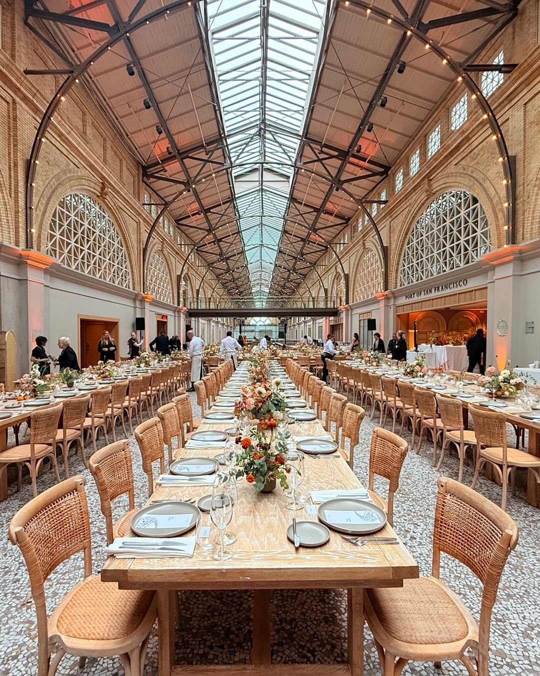 Long banquet tables set for a formal event inside a grand hall with a glass skylight ceiling.