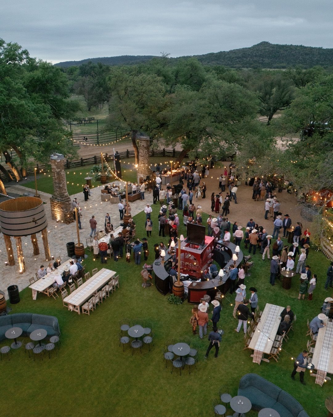 Aerial view of an outdoor wedding reception with guests, long tables, and string lights on a lawn.