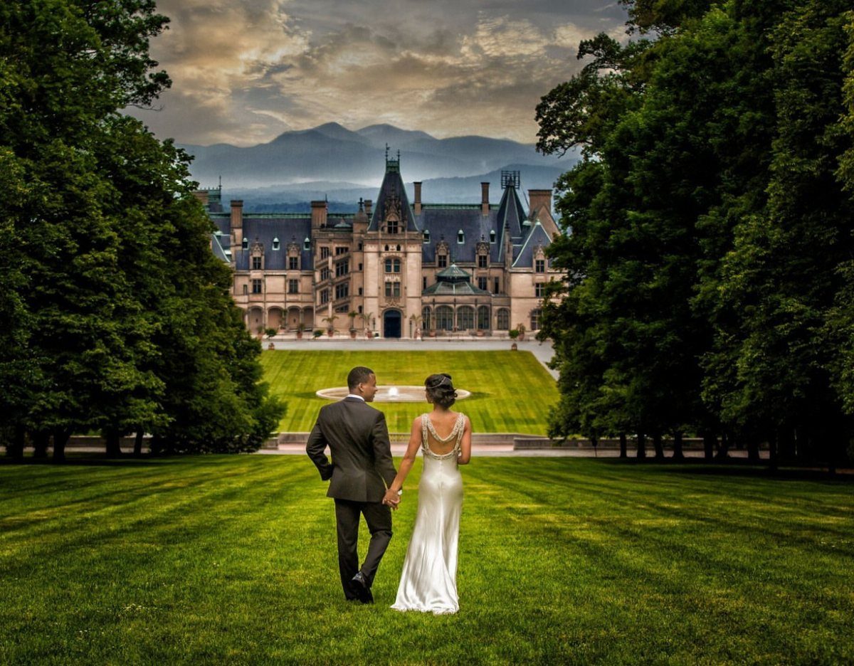 A bride and groom walking across a large lawn toward a grand historic mansion.