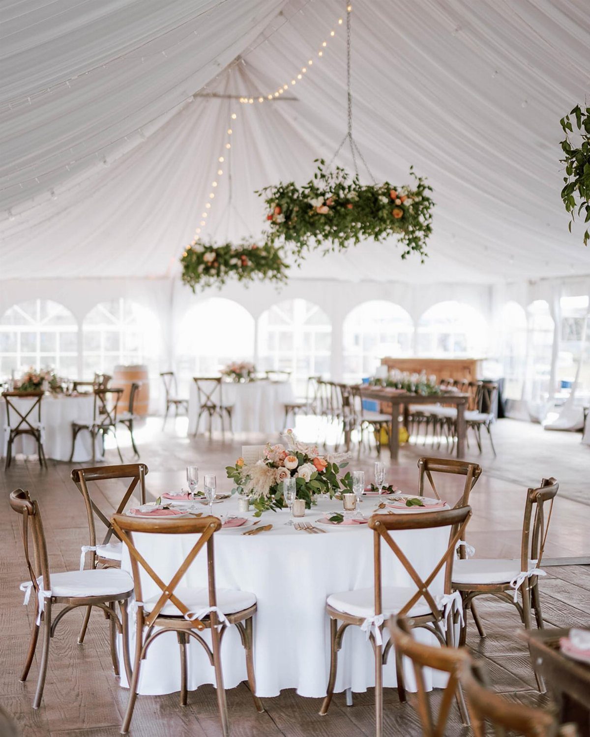 Elegant wedding reception tables with white linens and wooden chairs inside a large white tent.