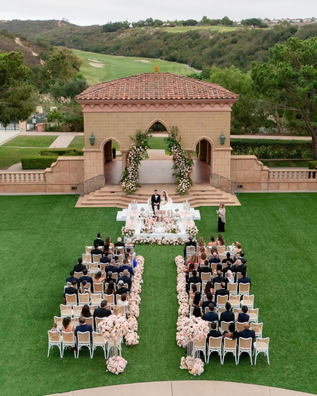 An outdoor wedding ceremony set on a green lawn with a stone pavilion and floral arrangements.