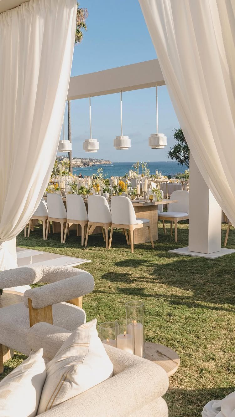 Outdoor dining table with white chairs set on grass overlooking the blue ocean.