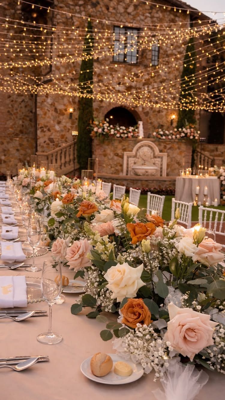 Long dining table decorated with peach and orange roses under hanging string lights by a stone building.