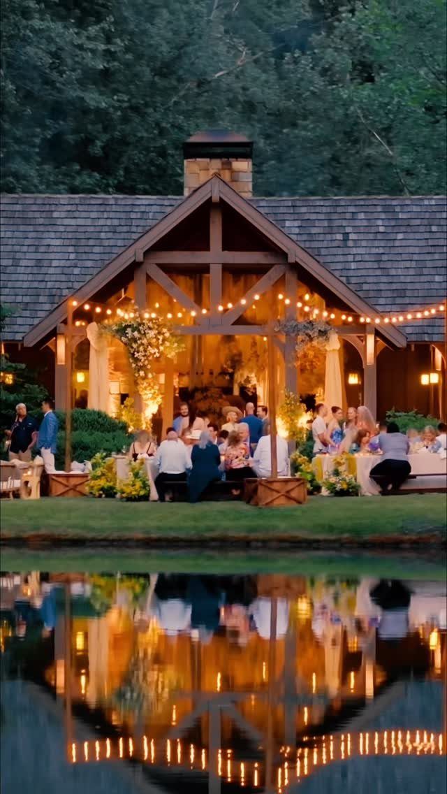 A lit wooden pavilion hosting a wedding dinner, reflected in a still pond at dusk.
