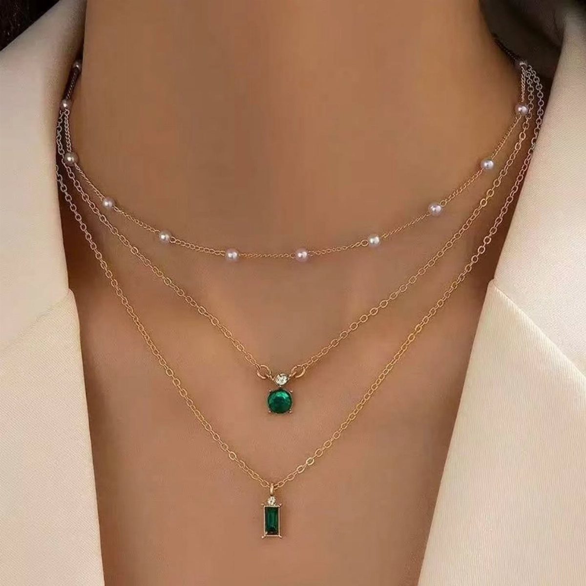 Three layered gold necklaces with pearls and green gemstone pendants worn on a person's neck.