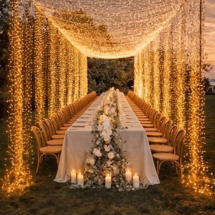 7 Long Table Wedding Reception Ideas for Outdoor Celebrations 4 A long wedding table decorated with white flowers and candles under a canopy of golden fairy lights.